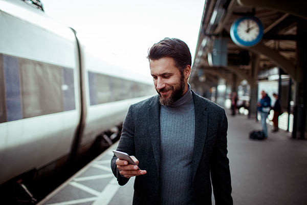 Hombre mirando y sonriendo a su teléfono mientras está en una estación de tren, caminando junto al tren.